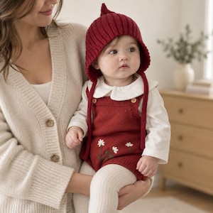 May include: A dark red knitted romper featuring white embroidered daisy flowers, worn with a matching ribbed bonnet with ties and a long-sleeved white shirt with a rounded collar, paired with cream-coloured ribbed leggings.
