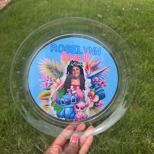 May include: A clear circular plastic plate featuring a custom graphic print. The design includes the text "ROSELYNN Turns 10" above an image of a young person wearing a floral crown, surrounded by tropical flowers, leaves, and the characters Stitch and Angel.