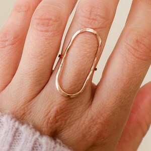 May include: A gold oval ring with a hammered texture. The ring is worn on the middle finger of a hand with nude nail polish.