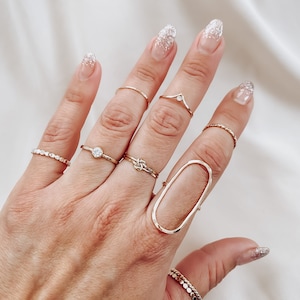 May include: A hand wearing a variety of gold rings, including a large oval ring, a knot ring, and a ring with a small stone. The nails are painted with a sparkly white polish.