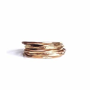 Puede incluir: Una pila de anillos finos de oro, cada uno con una textura martillada ligeramente diferente. Los anillos están apilados uno encima del otro, creando un aspecto único y moderno.