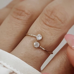 Op de afbeelding: Een gouden ring met twee kleine, heldere stenen op een dunne band. De ring wordt aan een vinger gedragen.
