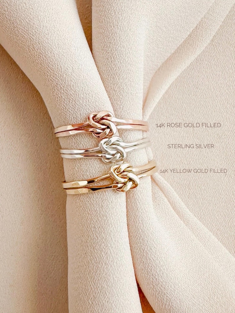 May include: Three gold rings with a knot design. The top ring is 14k rose gold filled, the middle ring is sterling silver, and the bottom ring is 14k yellow gold filled.