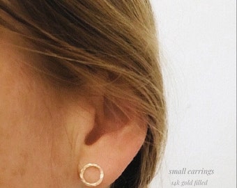 Pode incluir: Um par de pequenos brincos de argola de ouro preenchido. Os brincos são simples e delicados, com uma textura martelada. O texto "small earrings 14k gold filled" é visível na imagem.