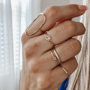 May include: A set of gold rings, including a large oval ring, a knot ring, and two thin bands. The rings are worn on a person's fingers.