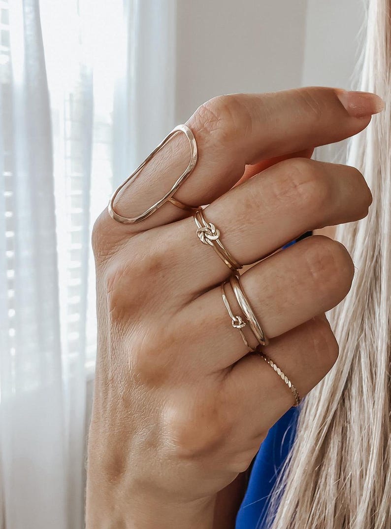 May include: A set of gold rings, including a large oval ring, a knot ring, and two thin bands. The rings are worn on a person's fingers.