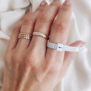 May include: A hand wearing three gold rings and a ring sizer tool. The ring sizer is white with black markings and the text "PAT. PEND. 91010853".