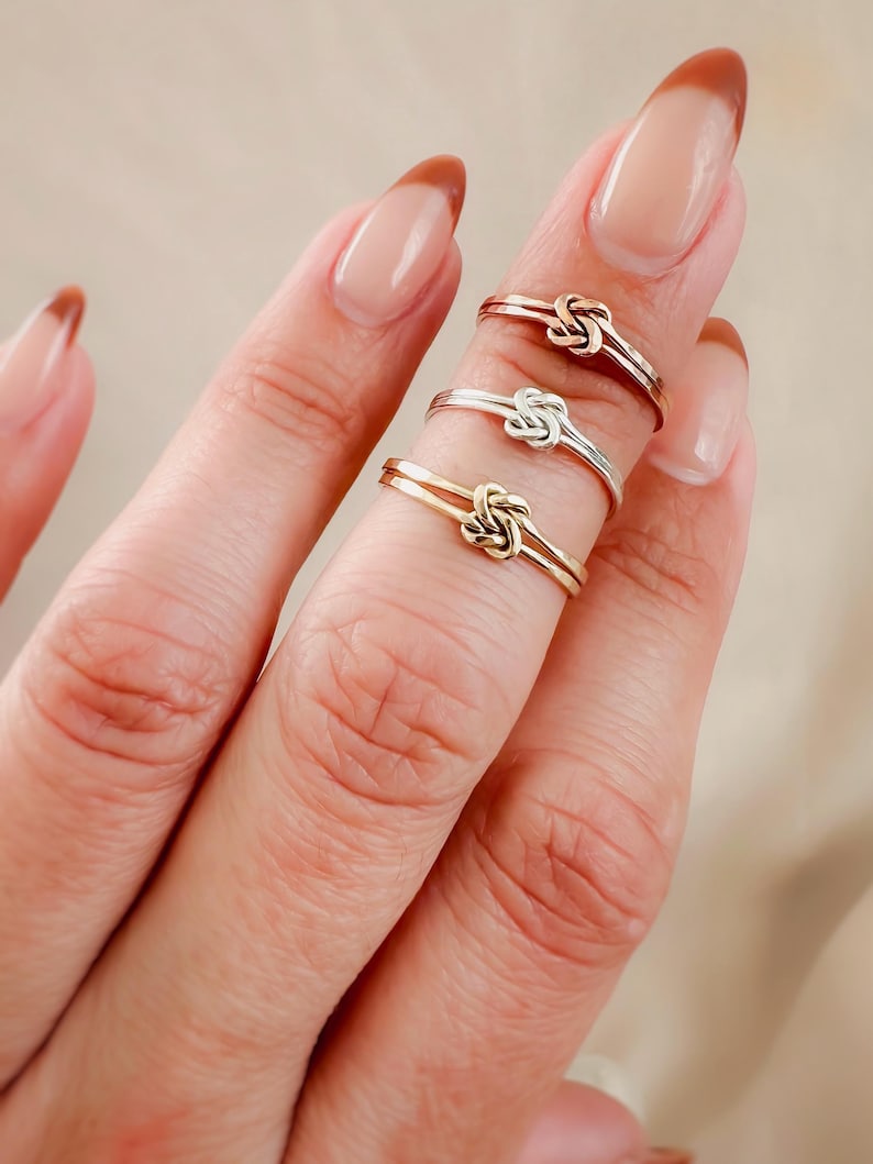 May include: Three gold, silver, and rose gold knot rings stacked on a finger. The rings are simple and delicate, with a small knot design.