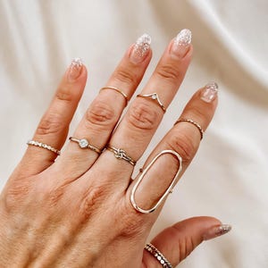 May include: A hand wearing multiple gold rings, including a large oval ring, a thin band with a small stone, a knot ring, and a delicate band with a small stone. The nails are painted with a white glitter polish.