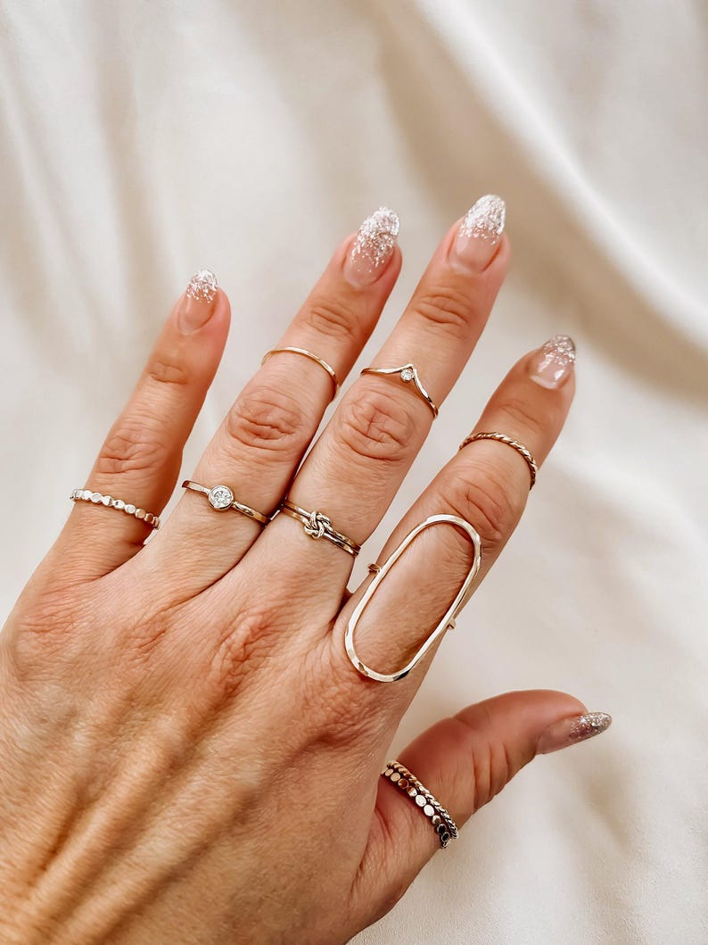 May include: A hand wearing multiple gold rings, including a large oval ring, a thin band with a small stone, a knot ring, and a delicate band with a small stone. The nails are painted with a white glitter polish.