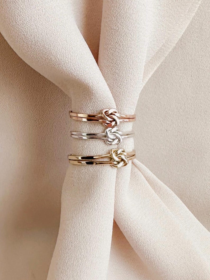 May include: Three gold, silver, and rose gold knot rings stacked on top of each other. The rings are all the same size and have a simple, elegant design.