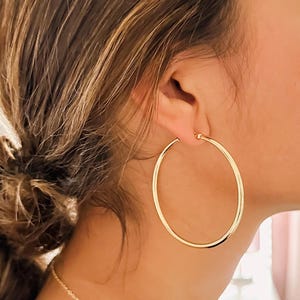 May include: Gold-colored hoop earrings. The earrings are large and circular, with a smooth, polished surface. A delicate gold necklace is also visible.