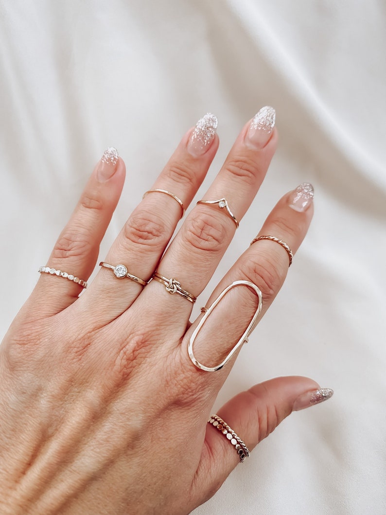 May include: A hand wearing a variety of gold rings, including a large oval ring, a knot ring, and a ring with a small stone. The nails are painted with a sparkly white polish.
