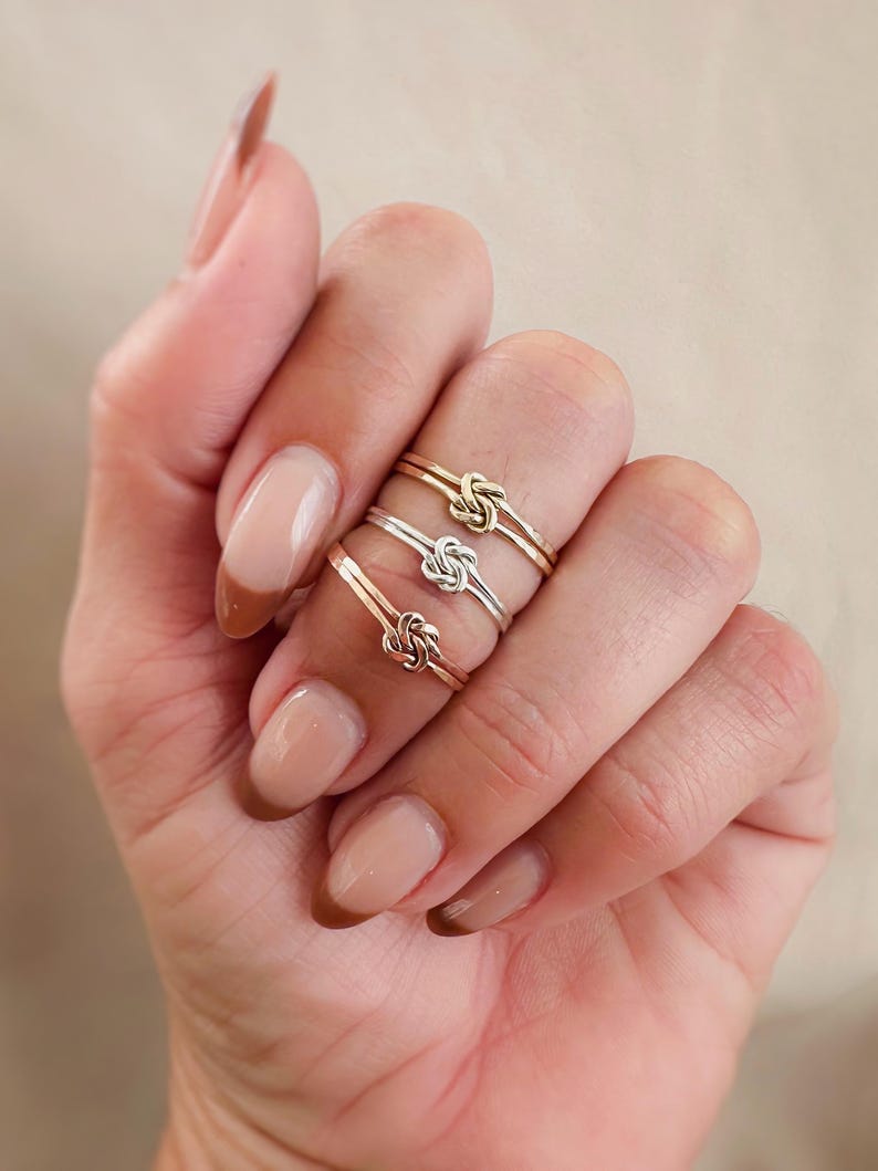 May include: Three gold, silver, and rose gold knot rings stacked on a finger.