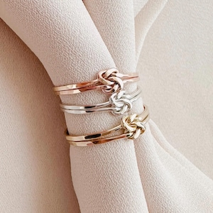 May include: Three gold rings with a knot design. The rings are stacked on top of each other. The top ring is rose gold, the middle ring is silver, and the bottom ring is yellow gold.