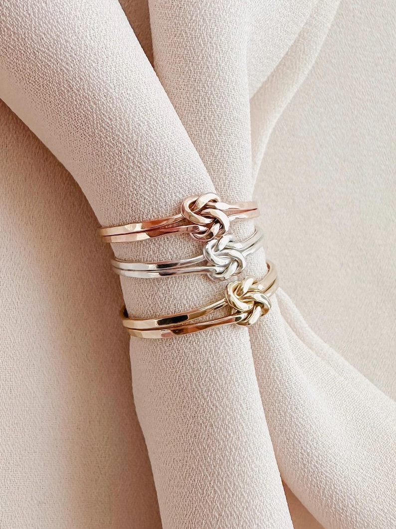 May include: Three gold rings with a knot design. The rings are stacked on top of each other. The top ring is rose gold, the middle ring is silver, and the bottom ring is yellow gold.