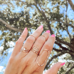 May include: A hand wearing a variety of gold rings, including a large oval ring, a ring with a small clear stone, and a ring with a delicate arch design.