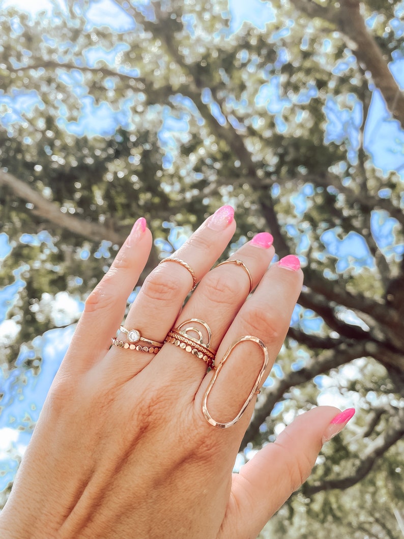 May include: A hand wearing a variety of gold rings, including a large oval ring, a ring with a small clear stone, and a ring with a delicate arch design.