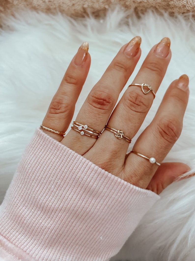 May include: A hand wearing a set of five gold rings. The rings include a simple band, a ring with three small stones, a knot ring, a ring with a pearl, and a heart-shaped ring.