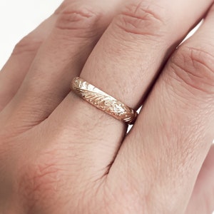 May include: A gold band ring with an intricate floral design. The ring is worn on a finger.