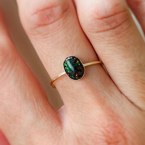 May include: A gold ring featuring an oval-shaped black opal gemstone with iridescent green and blue flashes. The opal is set in a delicate gold band, showcasing the stone's natural beauty. The ring is a piece of jewelry.