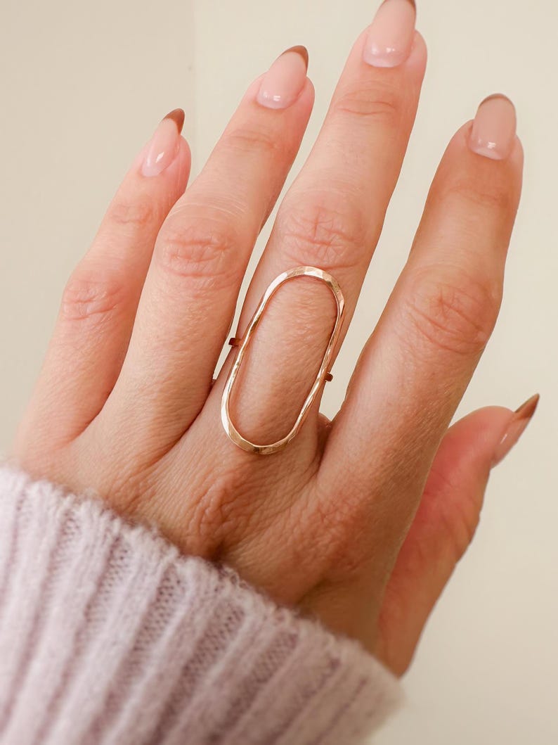 May include: A gold oval ring with a hammered texture. The ring is worn on the middle finger of a hand with nude nail polish.