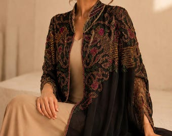 May include: A black open-front jacket featuring intricate gold, red, and green embroidery and beadwork, worn over a solid beige dress. The jacket has long sleeves with sheer panels and a high collar, showcasing detailed floral and scroll patterns.