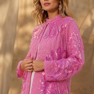 May include: A bright pink long-sleeved jacket featuring an intricate pattern of iridescent sequins and beadwork. The garment has a high neckline, a straight front opening, and a structured fit, showcasing detailed shell-like motifs across the fabric.