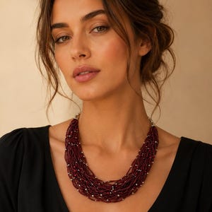 Op de afbeelding: Een meerstrengs ketting met talloze dunne draden van donkerrode kraaltjes, afgewisseld met kleine, reflecterende zilverkleurige metalen kralen. De ketting heeft een gelaagd, getextureerd uiterlijk en is afgewerkt met zilverkleurige eindkappen.