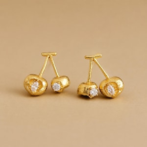 May include: A pair of gold-toned stud earrings shaped like cherries. Each earring features two textured, rounded fruit forms connected by thin stems. A single clear, faceted gemstone is embedded in the center of each cherry-shaped element.