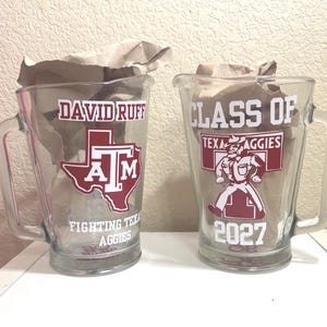 May include: Two clear glass pitchers with handles, each filled with crumpled brown paper. The left pitcher features the text DAVID RUFF, a maroon Texas A&M logo, and FIGHTING TEXAS AGGIES. The right pitcher displays CLASS OF, a maroon mascot, and 2027.