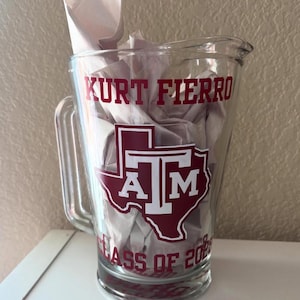 May include: A clear glass pitcher featuring the maroon and white Texas A&M logo, the name KURT FIERRO, and the text CLASS OF 2028. The pitcher has a sturdy handle and a pouring spout, with white paper filler visible inside the glass container.