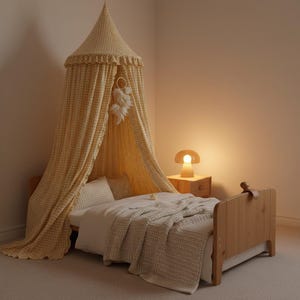 May include: A yellow and white gingham bed canopy with a ruffled top edge hangs over a wooden bed frame. The bed is dressed in white bedding with a textured waffle-weave blanket. A small mushroom-shaped lamp sits on a wooden nightstand nearby.