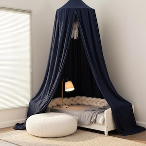 Könnte beinhalten: Ein dunkelblauer Stoffhimmel hängt von der Decke über einem weißen Bettgestell. Das Bett verfügt über einen cremefarbenen geflochtenen Bettschutz, eine hellgraue Decke und eine goldene Lampe. Ein runder, strukturierter weißer Hocker steht auf einem beigen Teppich.