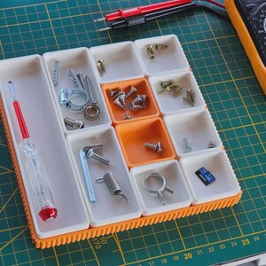 Pode incluir: Um organizador de armazenamento modular com compartimentos de plástico brancos e laranja contendo vários parafusos metálicos, porcas, uma mola, uma braçadeira, um pequeno cartão de memória e uma chave de fenda com cabo vermelho.