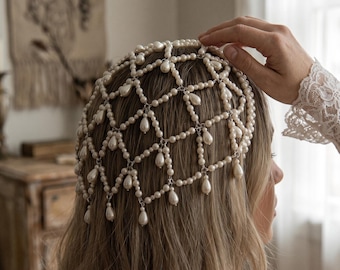May include: A headpiece constructed from a lattice of white spherical pearls and dangling teardrop-shaped pearls. The accessory features a net-like design that covers the crown of the head, with silver-toned metal links connecting the pearl strands.