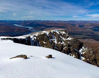 May include: A high-altitude view of a snow-covered mountain peak with exposed dark rocky cliffs. In the distance, a long, narrow body of water stretches through a vast landscape of brown hills under a bright blue sky with scattered white clouds.
