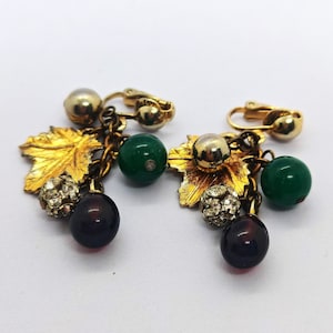 May include: A pair of gold-tone clip-on earrings featuring dangling charms shaped like grape clusters and leaves. Each earring includes a textured gold leaf, a smooth green bead, a dark red bead, a gold sphere, and a small crystal-encrusted ball.