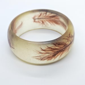 Può includere: Un bracciale rigido circolare traslucido realizzato in un materiale trasparente simile alla resina. Il bracciale presenta piume marroni incorporate visibili in tutta la struttura, creando un aspetto organico e stratificato all'interno della fascia liscia.