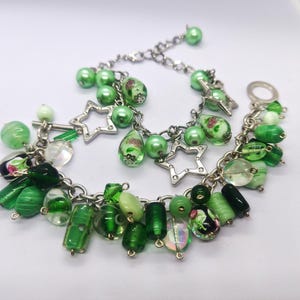 May include: A charm bracelet featuring a silver-toned chain adorned with various green beads, including translucent glass, opaque spheres, and floral-patterned lampwork pieces, interspersed with open silver-toned star charms and a toggle clasp.