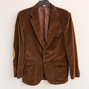May include: A brown corduroy blazer featuring a notched lapel, two front buttons, two flap pockets, and tan elbow patches. The sleeves are finished with three small buttons at each cuff, and the interior is lined with a smooth brown fabric.