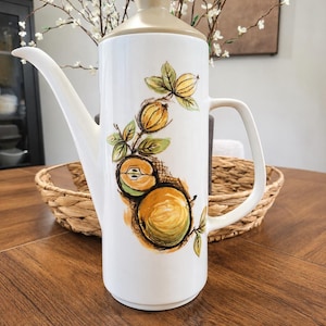 Puede incluir: Una cafetera alta de cerámica blanca con una ilustración pintada de frutas amarillas y verdes con hojas en la parte frontal. La cafetera tiene un pico largo y curvado, un asa lateral y una tapa de color tostado claro a juego con un pomo.