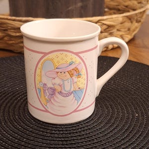 Könnte beinhalten: Ein weißer Keramikbecher mit einer zentralen ovalen Illustration einer Figur mit geflochtenem Haar, die einen breitkrempigen rosa Hut und ein rosa Kleid trägt. Das Design ist von rosa Linien und kleinen Punkten umrahmt.