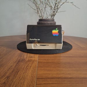 Puede incluir: Una caja de almacenamiento Flip'n File/25 con base beige y tapa translúcida oscura. La tapa presenta una pegatina del logotipo de Apple con los colores del arcoíris. La parte frontal de la base beige muestra el texto Flip'n File/25 y un icono.