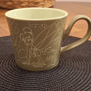 Puede incluir: Una taza de cerámica color verde salvia con una ilustración de líneas blancas de Campanilla con alas y patrones de estrellas giratorias. La taza tiene un acabado liso, un borde ancho y un asa curva en el lado derecho del diseño.