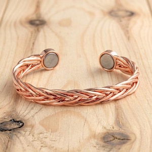 Pode incluir: Uma pulseira tipo bracelete de cor cobre com um design trançado de vários fios. A banda aberta termina em duas extremidades magnéticas circulares com um acabamento metálico prateado liso nas faces interiores dos terminais circulares.