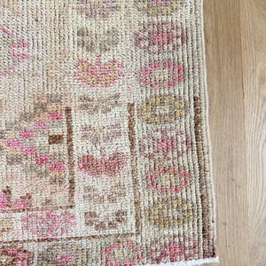May include: A hand-knotted wool rug featuring a cream-colored base with intricate geometric patterns in shades of soft pink, muted brown, and beige. The design includes repeating floral-like motifs and a structured border with oval shapes.