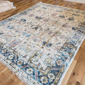May include: A rectangular area rug featuring a cream-colored field with an intricate pattern of floral and geometric motifs in shades of blue, muted pink, and gold. The rug has a wide, detailed border and short fringe along the edges.