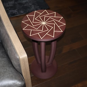 May include: A round side table with a deep maroon finish featuring a geometric pattern of light-colored triangles on the tabletop. The table is supported by three curved, vertical legs that connect to a circular base, all in a matching maroon color.