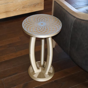 May include: A round side table with a metallic gold finish featuring a geometric light blue pattern on the top surface. The table has a circular base and three curved legs that connect to the underside of the tabletop in a central column design.